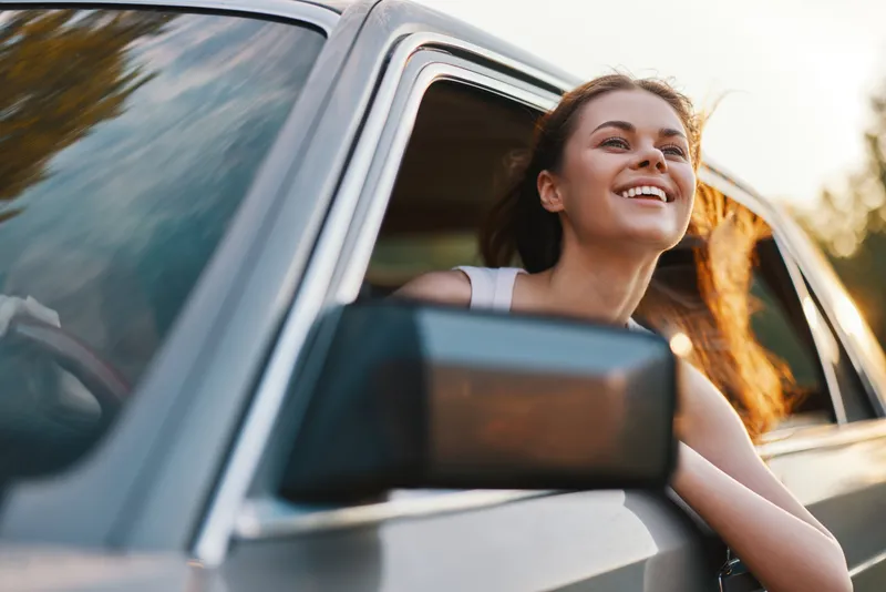 Young woman enjoying a drive with a joyful smile, hair blowing in the wind, summer vibes, outdoor adventure, and carefree emotions.