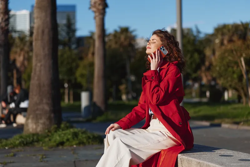 A woman in a red coat sits on a concrete bench in a sunny park, talking on a mobile phone with a thoughtful expression and relaxed posture.