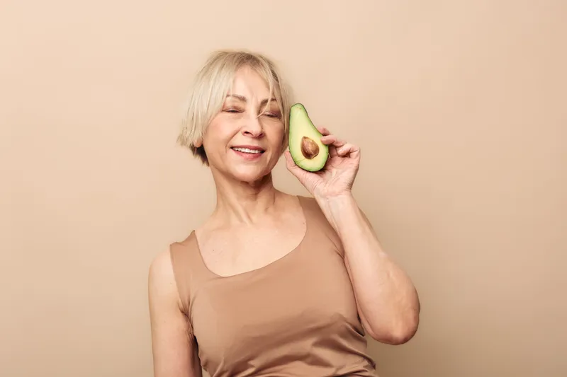 Smiling mature woman with short blonde hair holds half an avocado near her face on beige background. Concept of healthy lifestyle, nutrition, natural food, and wellness for seniors.