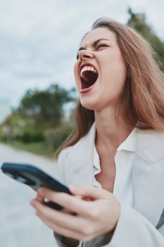 Frustrated woman holding smartphone shouting outdoors expressing anger and stress in casual white jacket with blurred nature background