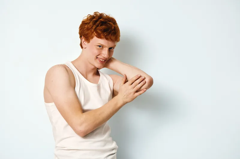 Young man with red hair and pale skin wearing white sleeveless shirt touching and examining his elbow against plain light background. Smiling with casual pose in studio lighting.