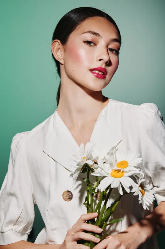 Portrait of a young woman with sleek dark hair wearing a stylish white blouse holding fresh daisies. Studio shot on pale green background. Concept of beauty, fashion, and creativity.