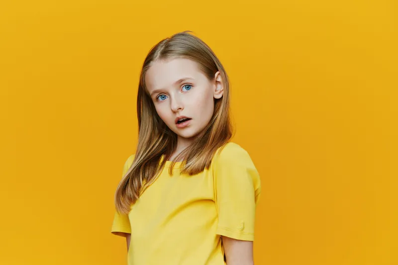 A young girl with blue eyes, wearing a bright yellow shirt, expressing curiosity and surprise against a vivid yellow background, showcasing playful childhood emotions