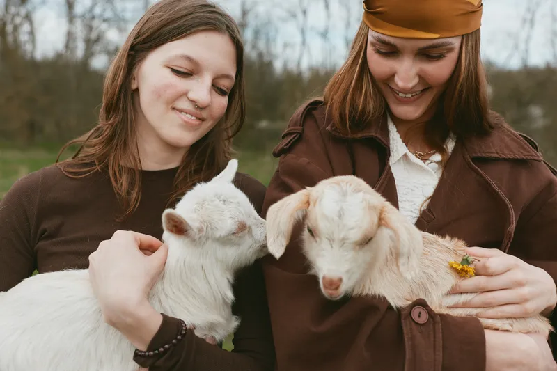 Two young women enjoy a peaceful moment holding baby goats outdoors in casual autumn clothing, smiling gently in a natural lifestyle setting.