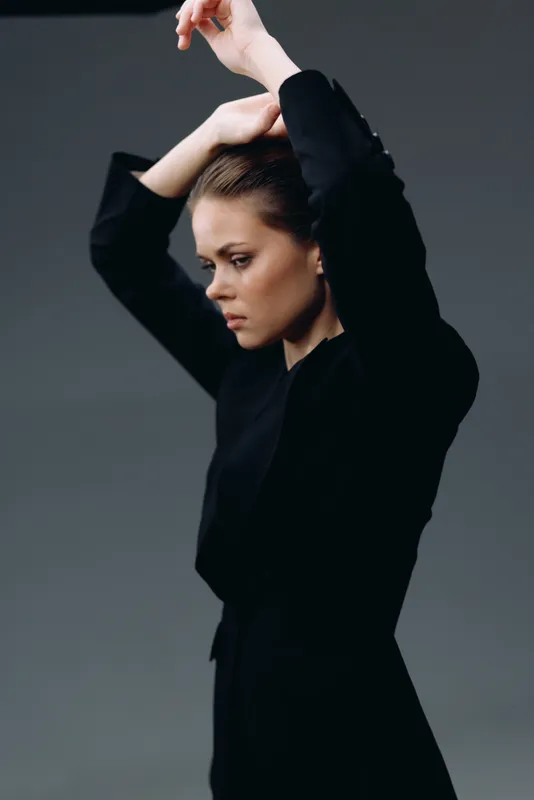 thoughtful woman in black clothing poses with raised arms against gray background, elegant style, focused expression