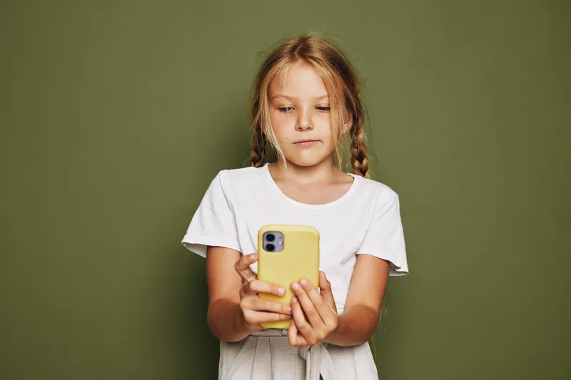 Young girl with braided hair wearing a white t-shirt looks focused while holding a yellow smartphone on a green background. Concept for technology, childhood, and communication.