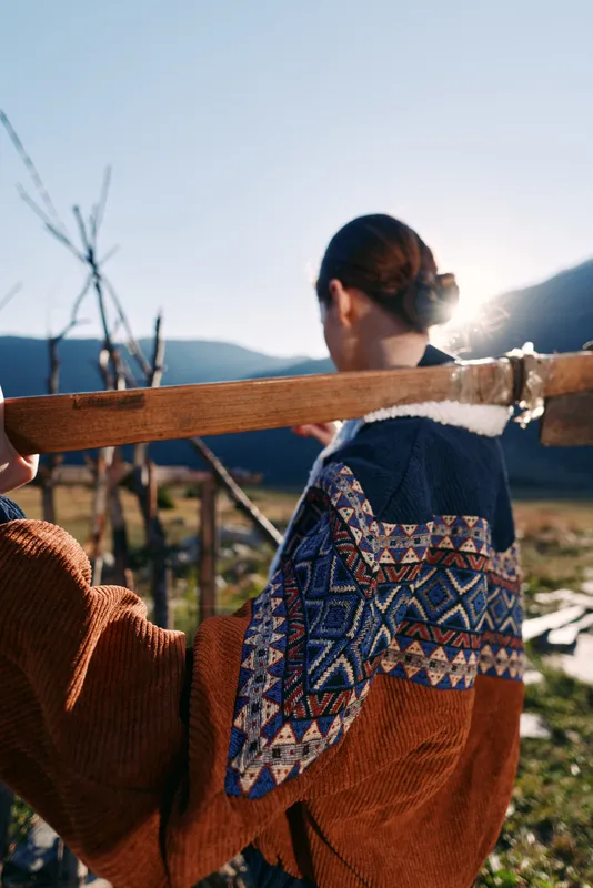 Woman back in patterned sweater carrying a wooden beam across a rural field at sunset with sun flare, rustic fence and cozy clothing for countryside lifestyle and outdoor work.