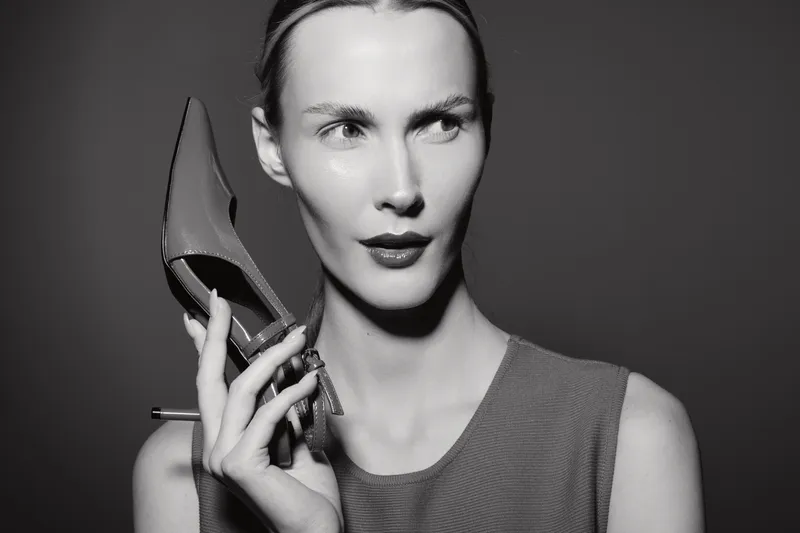 A slim white woman with light skin holds a high heel shoe near her face with a thoughtful expression. Black and white studio portrait with dramatic lighting and fashion concept.