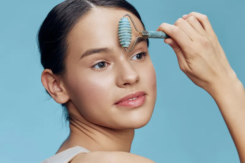 Young woman applying skincare with a jade roller on her forehead, showcasing a serene expression against a light blue background, emphasizing beauty and self care routines