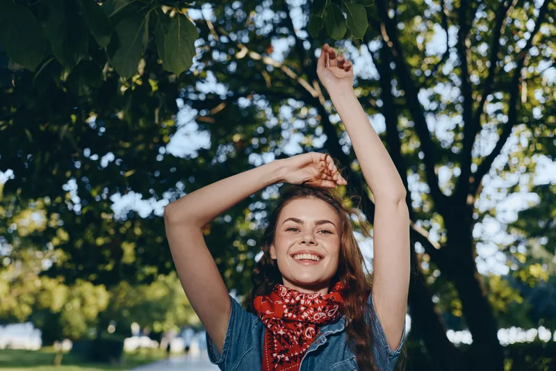 Lifestyle woman in modern boho-western denim street style with a red bandana smiles joyfully under green tree leaves in warm natural light, film color ambiance.