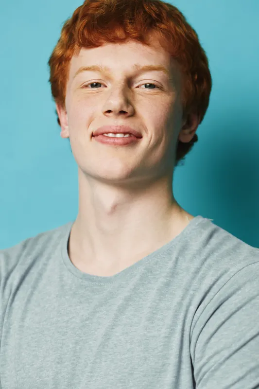Young man with red hair and freckles wearing gray t shirt smiles confidently against blue background portrait closeup casual studio lighting expression.