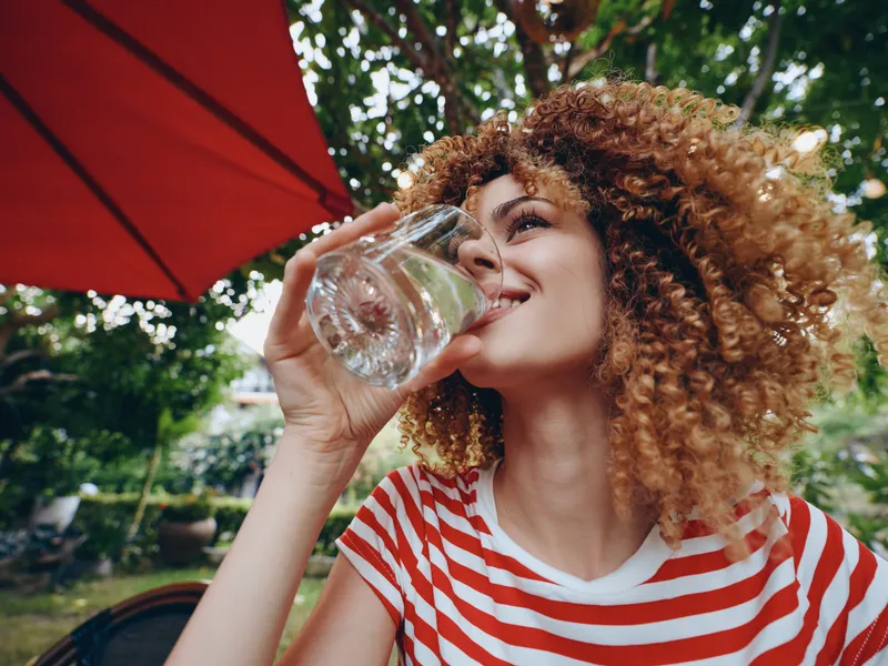 A woman with curly hair sips water from a glass outdoors, wearing a red and white striped shirt under a sunny patio setting.