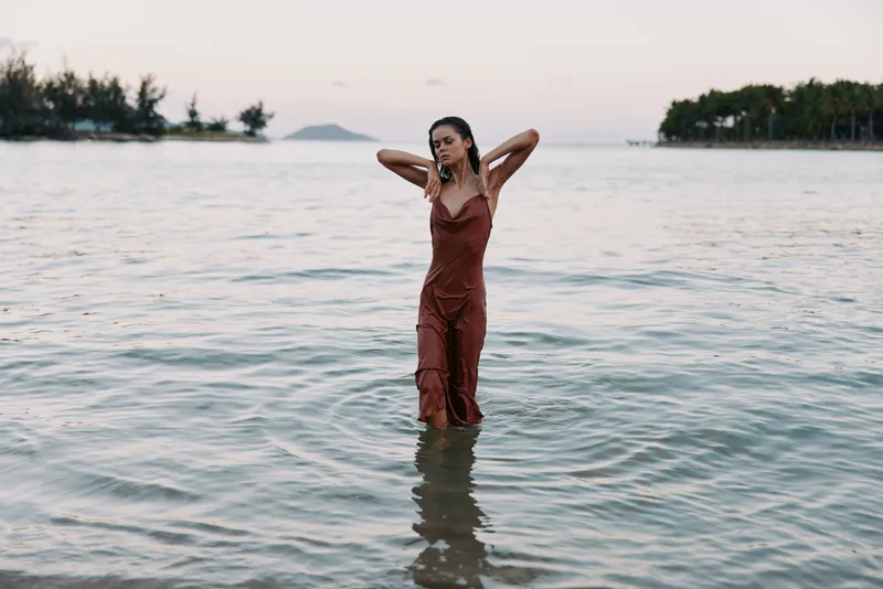 A solitary woman stands waist deep in calm sea water wearing a red dress, posing with arms raised behind her head, soft sunset light, tranquil coastal scene, reflective surface