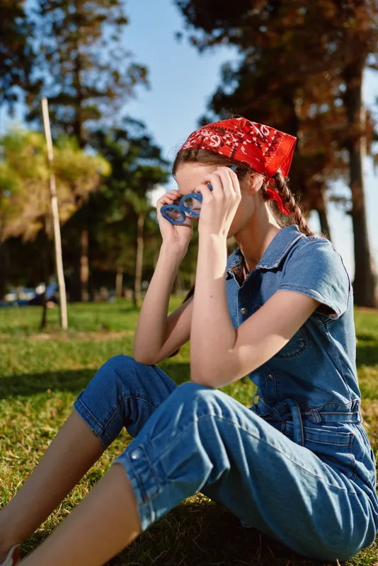 lifestyle woman in modern boho-western denim street style sitting on grass with red bandana and film color, enjoying outdoor sunny day in casual outfit