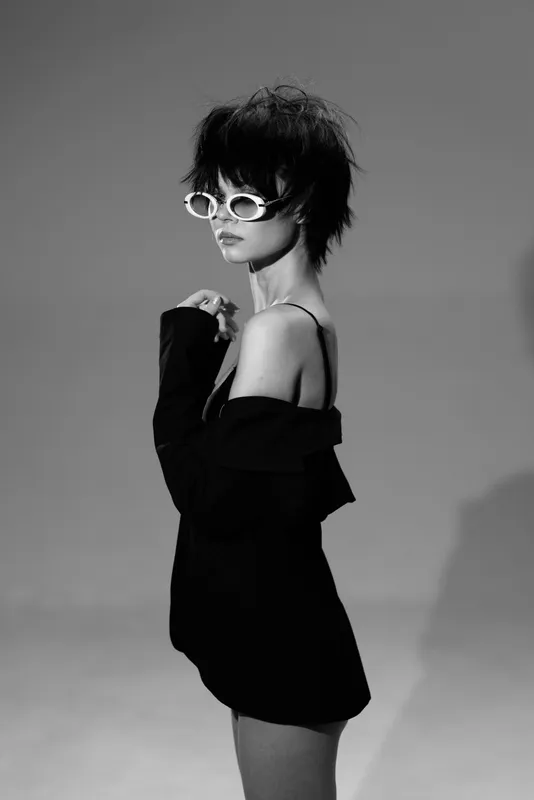 Black and white portrait of stylish woman wearing sunglasses and off-shoulder dress with modern hairstyle and confident pose.