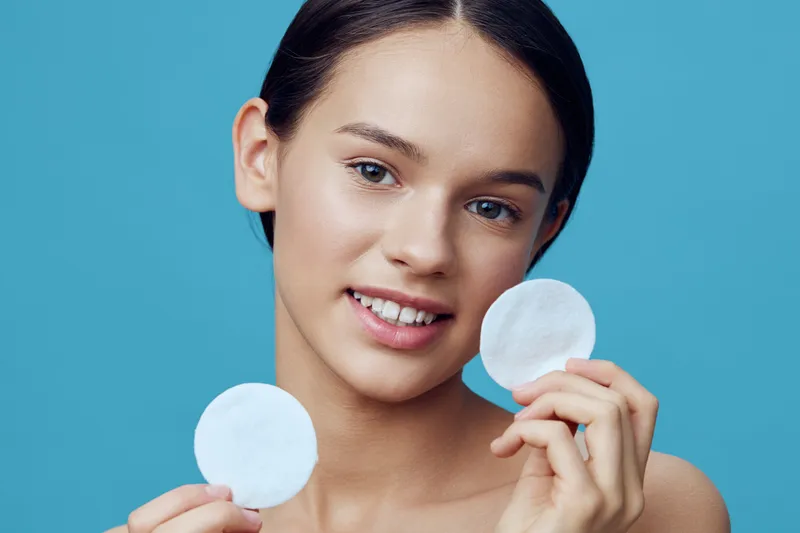 Young woman with clear skin holding facial cotton pads, smiling against a bright blue background, promoting skincare and beauty routines