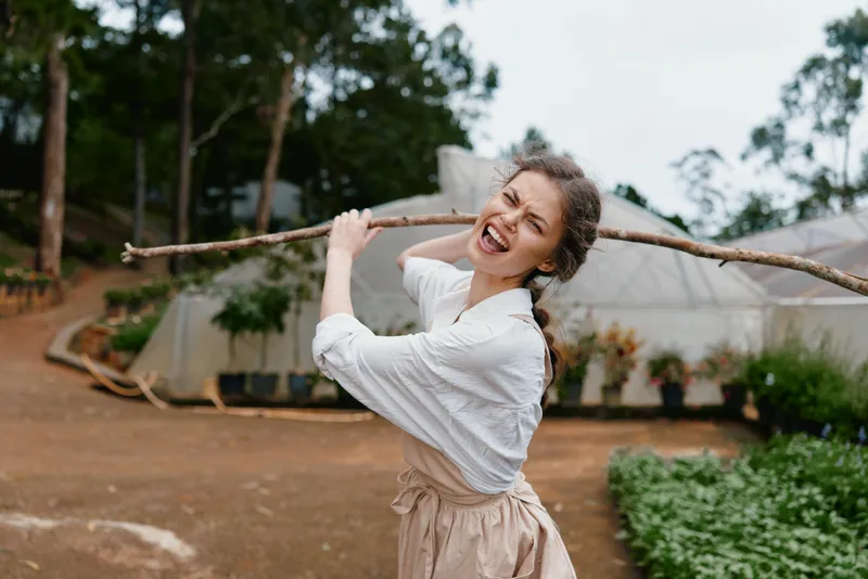 Cheerful young woman in an apron playfully holding a stick in a garden setting with greenery, showcasing joy and connection to nature