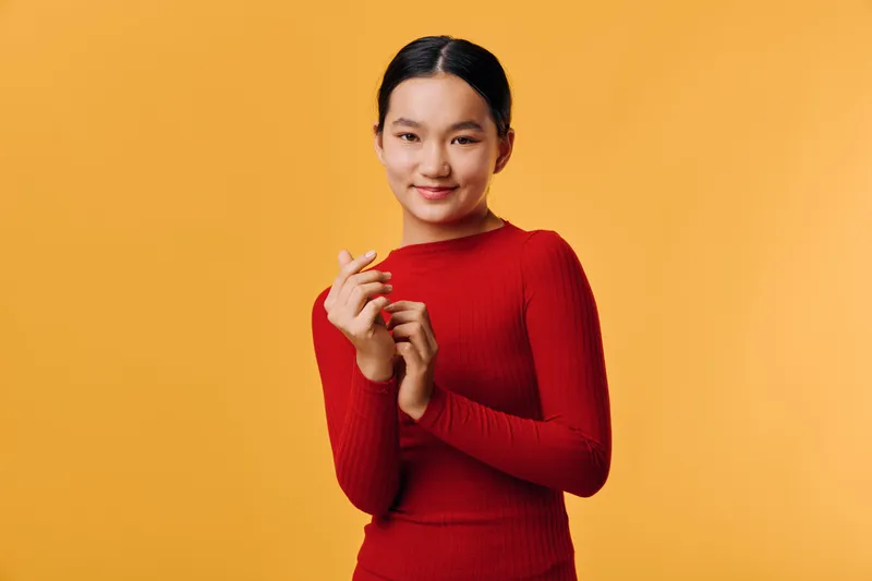 Young woman in red sweater standing on yellow background with hands close to face, smiling softly and looking at camera, casual portrait with minimal style and natural lighting.