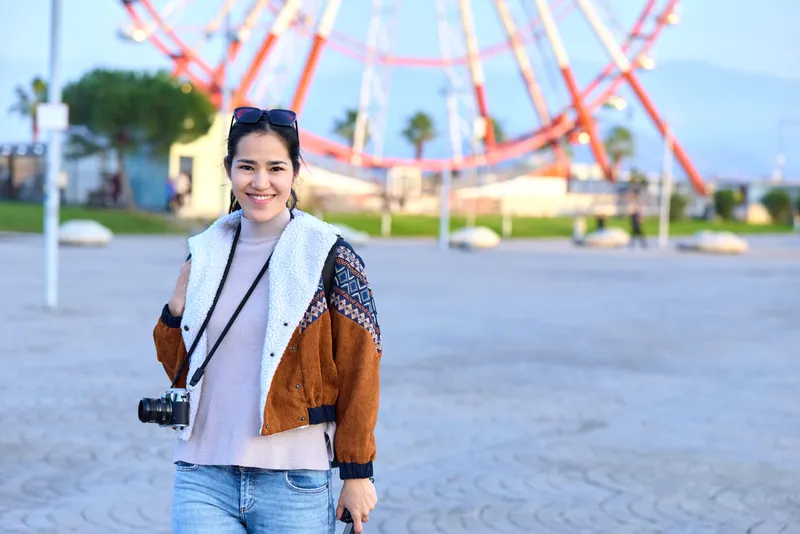 Woman with camera in a casual jacket stands in an outdoor amusement park, rollercoaster in the background, sunny day, capturing travel moments and street style.