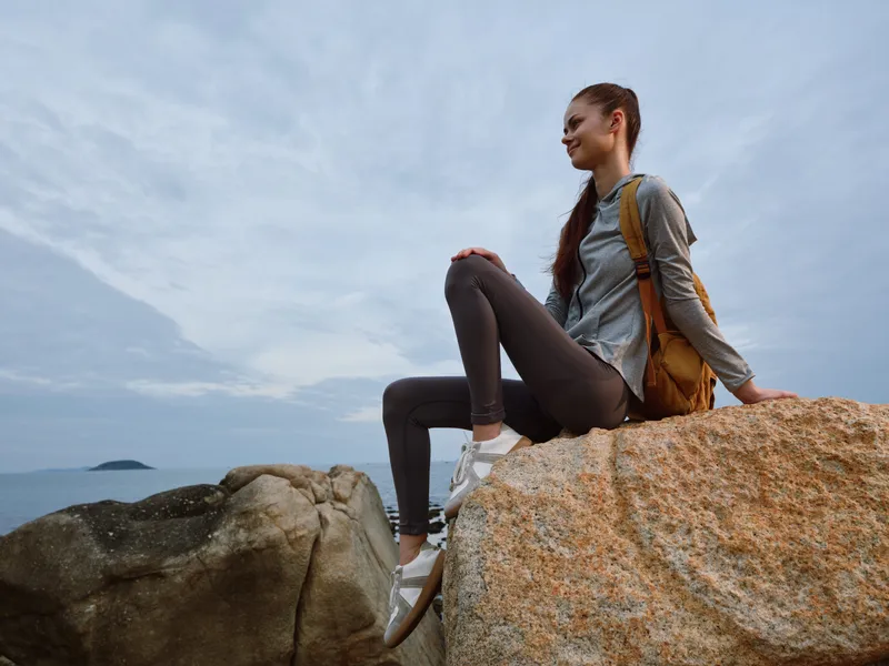 Young traveler enjoying nature, sitting on rocks by the sea, dressed in casual outdoor clothing, exuding a sense of peace and adventure