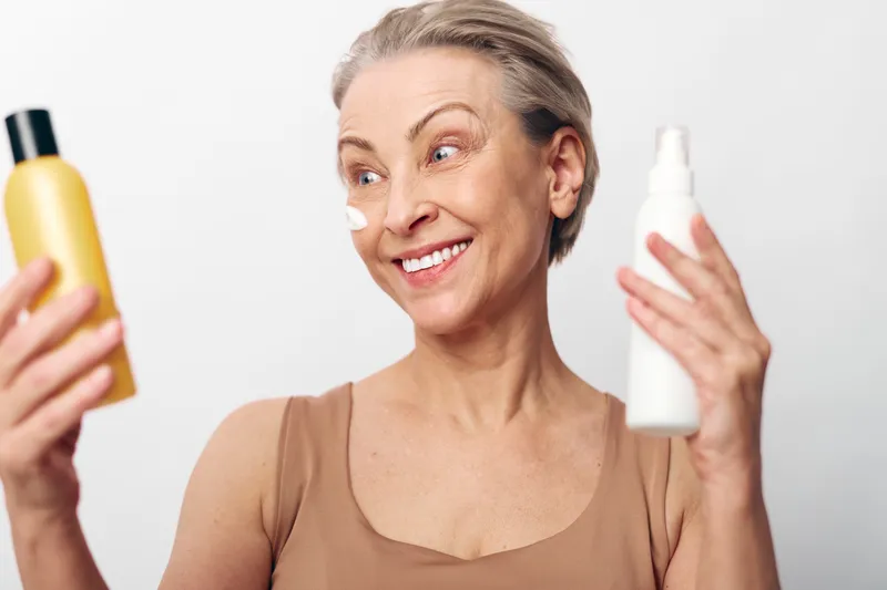 Smiling mature woman with short hair holds two skincare bottles and applies cream on cheek. White background, concept of beauty routine, facial care, and healthy skin for older adults.