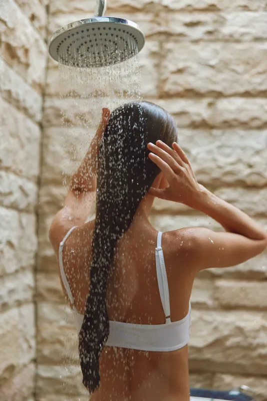 woman showering under outdoor rain shower with wet long hair and white bikini top against textured stone wall, relaxing summer scene, natural light