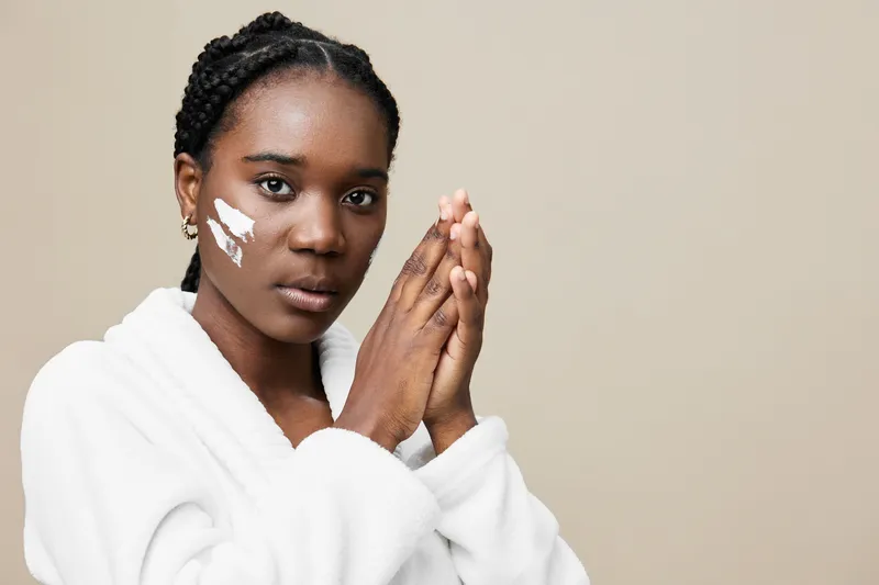 African woman applying skincare product, showcasing natural beauty and self care, wearing a cozy white robe, expressing tranquility against a neutral background