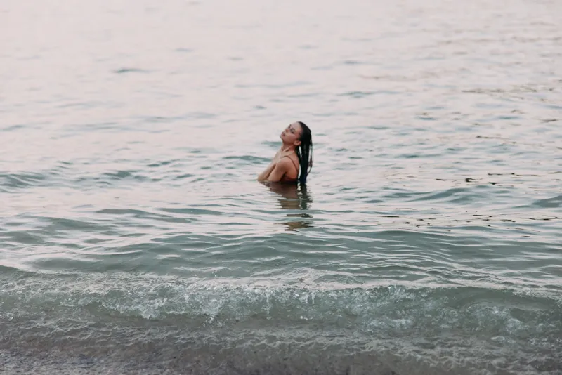 A lone woman stands waist deep in calm sea water, facing slightly upward as soft sunlight warms the skin, with gentle waves embracing the shoreline.