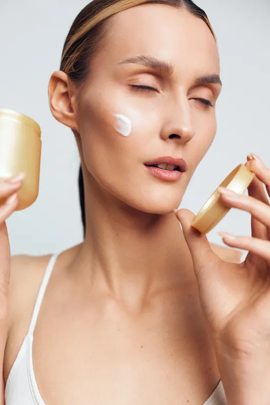 Young woman applies face cream on cheek skin holding cosmetic jar with closed eyes in white tank top isolated on light background for daily skincare routine and beauty care concept.