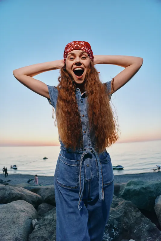 lifestyle woman in modern boho-western denim street style with long hair and red bandana smiling outdoors near sea at sunset in film color mood
