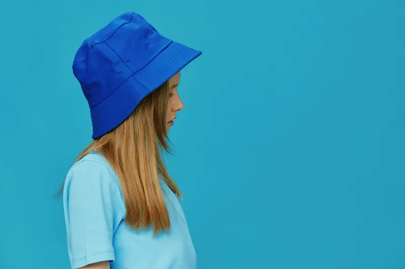 A young girl in a blue bucket hat and matching t shirt stands against a vibrant turquoise background, showcasing a playful and trendy summer vibe