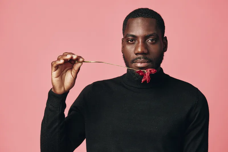 Creative conceptual portrait of African man in black turtleneck holding red chili pepper near mouth on pink background. Visual style with focus on expression and color contrast.