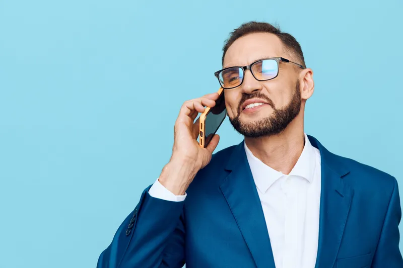 A professional businessman stands on a solid blue background, wearing a sharp suit and glasses, focused and confident as he talks on a mobile phone.