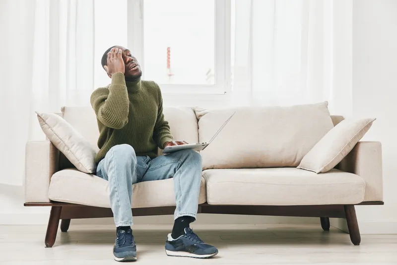 Frustrated young African man sitting on a couch with a laptop, expressing stress and anxiety about work or studies in a modern, bright living room environment