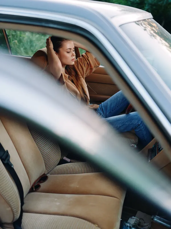 A young woman in a casual outfit relaxes inside a vintage car, expressing a sense of tranquility amidst nature's greenery.