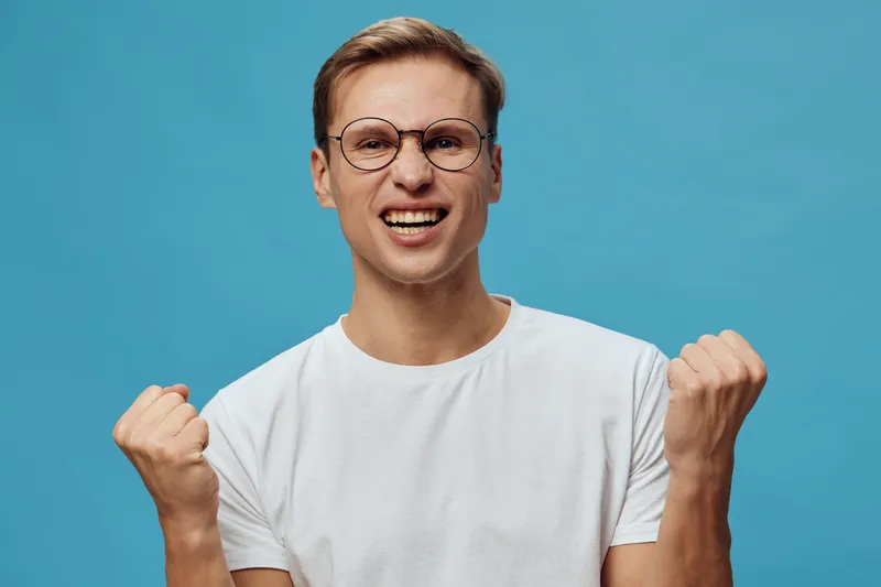 Smiling woman with short light brown hair, wearing glasses and a white t-shirt, showing fists in excitement, studio shot with solid bright blue background, cheerful mood, positive emotion, casual styl