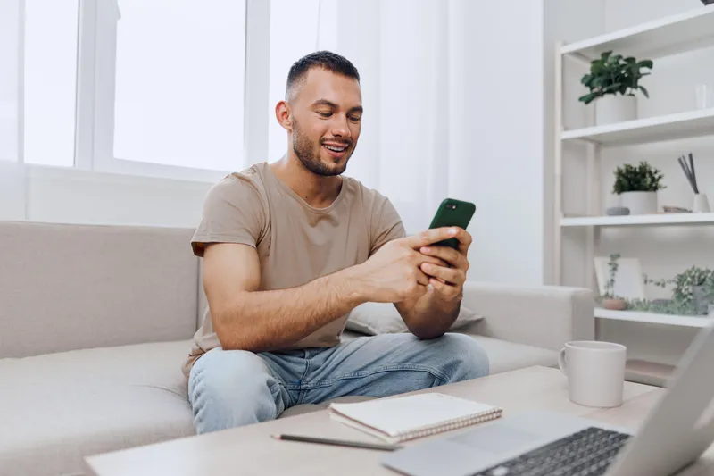 Smiling man using smartphone in modern living room, enjoying connectivity and comfort. A relaxed atmosphere with a cozy interior enhances the experience of technology.