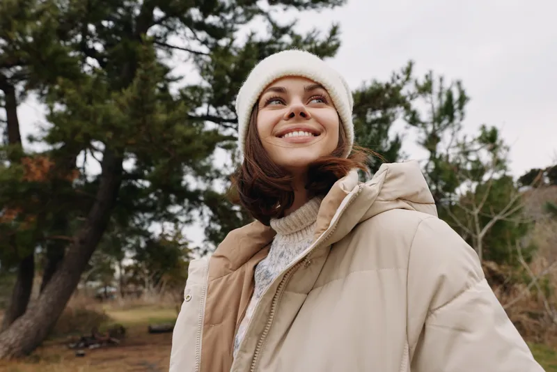Outdoor woman wearing a beige jacket and white beanie, smiling up at the trees in a park setting, showcasing casual fashion and fresh air style.