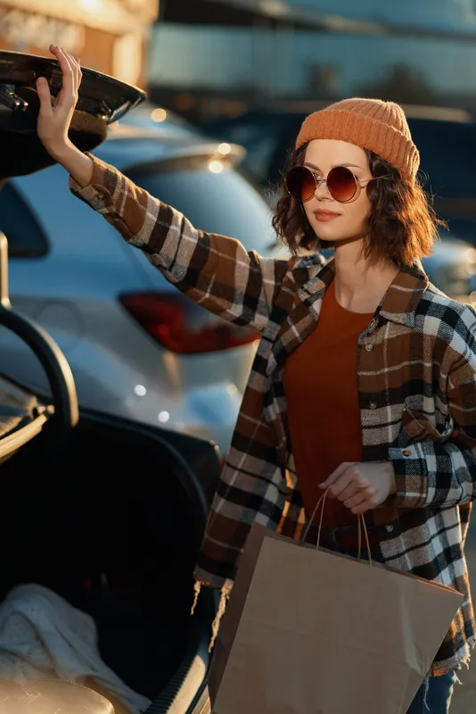 Woman shopping at car trunk with sunglasses and beanie, plaid coat and paperbag in golden hour glow, candid lifestyle portrait emphasizing authenticity, mindful living and emotional storytelling