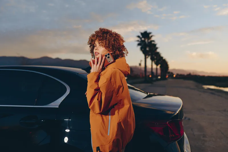 Woman lifestyle outdoors at sunset near car, holding a phone to her ear, curly hair and warm brown jacket, relaxed pose by the trunk, soft evening light, quiet street background