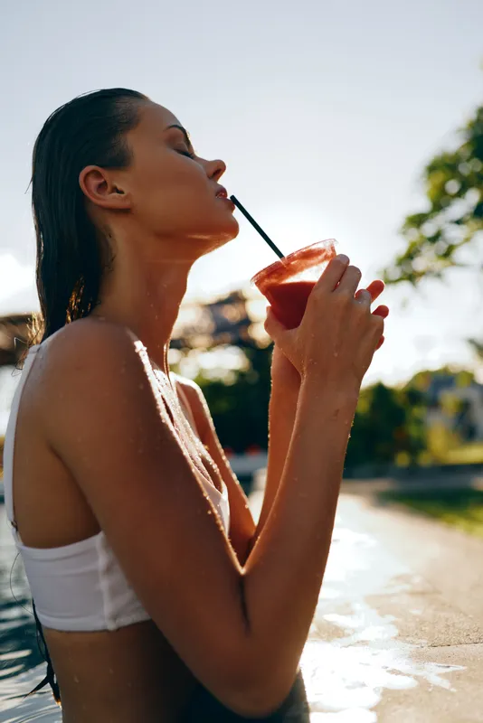 Summer woman by the pool enjoys a refreshing drink, wearing a white bikini top, hair wet, sipping through a straw, bright sunlight and relaxed mood in an outdoor leisure moment.