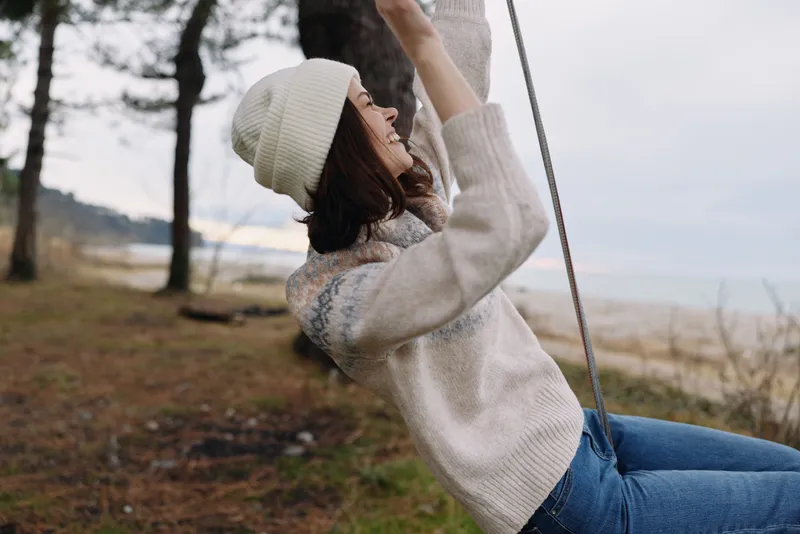 Woman on a swing outdoors wearing a knit sweater and beanie, enjoying a breezy moment by a beachside park with trees, calm air, and a relaxed, carefree mood.