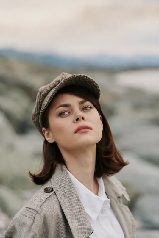 Young woman with brown hair wearing beige coat and cap looks thoughtfully outdoors with blurred natural background. Concept of autumn fashion, contemplation, and serene mood.