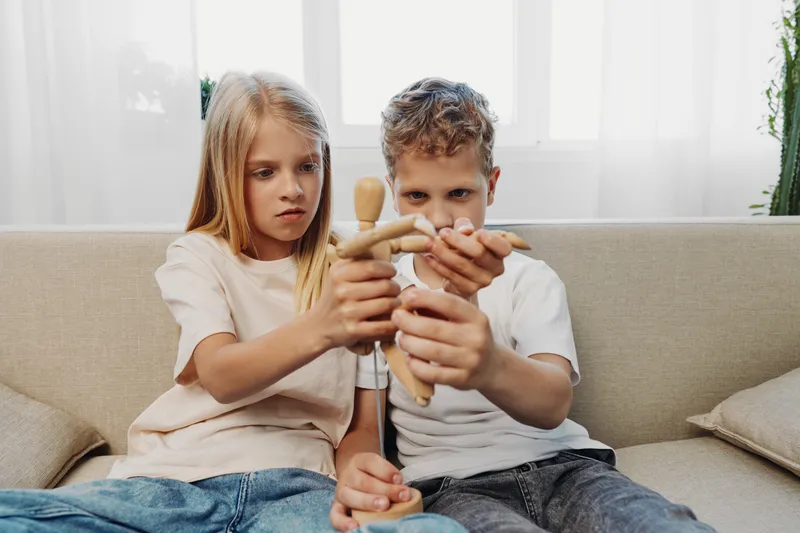 Boy and girl sitting on sofa play with wooden mannequin, focusing on creativity and imagination in a cozy home environment with natural light from window behind.