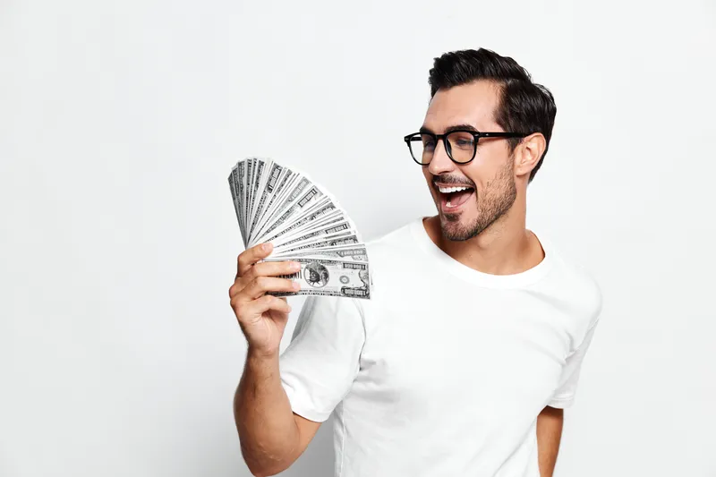 Smiling man wearing glasses holds a fan of money while looking at the camera, showcasing excitement and joy on a plain white background. Finance concept