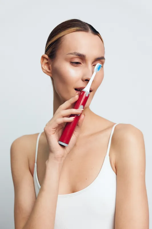 A young woman in a white tank top holds a red electric toothbrush near her face, preparing for oral hygiene. The clean white background emphasizes dental care and personal grooming.