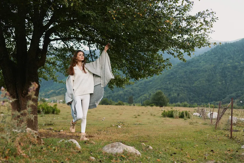 A woman enjoys outdoor life in a peaceful countryside field, carrying a blanket under a leafy tree. Bright natural light highlights casual clothing and a relaxed mood.