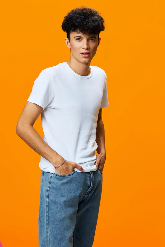 young man in white t-shirt and blue jeans posing confidently with curly hair against vibrant orange background, casual style, modern fashion look, studio portrait