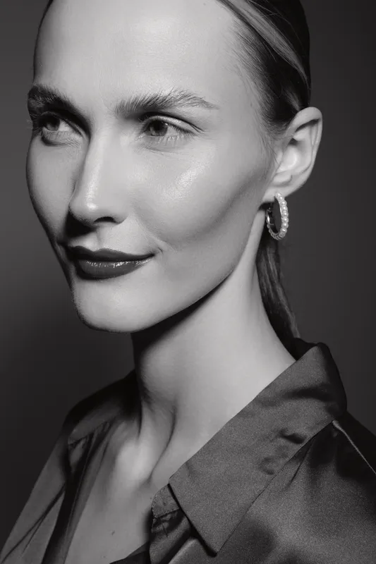Close-up portrait of a young white woman with fair skin and slicked back hair wearing a satin shirt and hoop earrings. Studio lighting highlights her smooth complexion and elegant look.