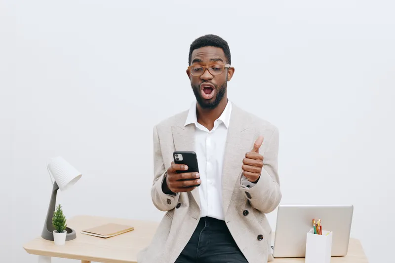Confident young African American man in formal beige blazer, glasses, using mobile phone with surprised expression, gesturing thumbs-up, standing in modern office with desk and accessories. People lif
